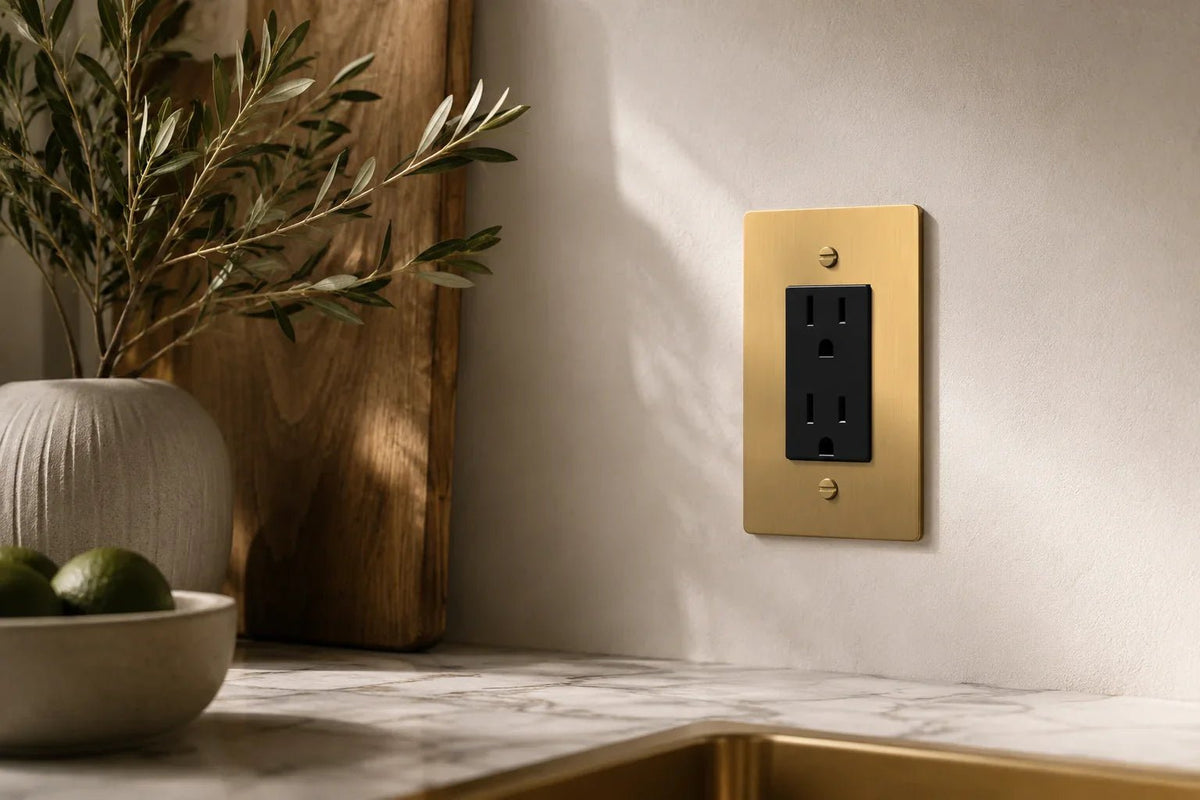 Luxury solid brass electrical outlet cover with a black duplex receptacle installed on a textured wall in a modern kitchen. Premium decorative brass outlet plate with warm natural lighting and high-end interior design.